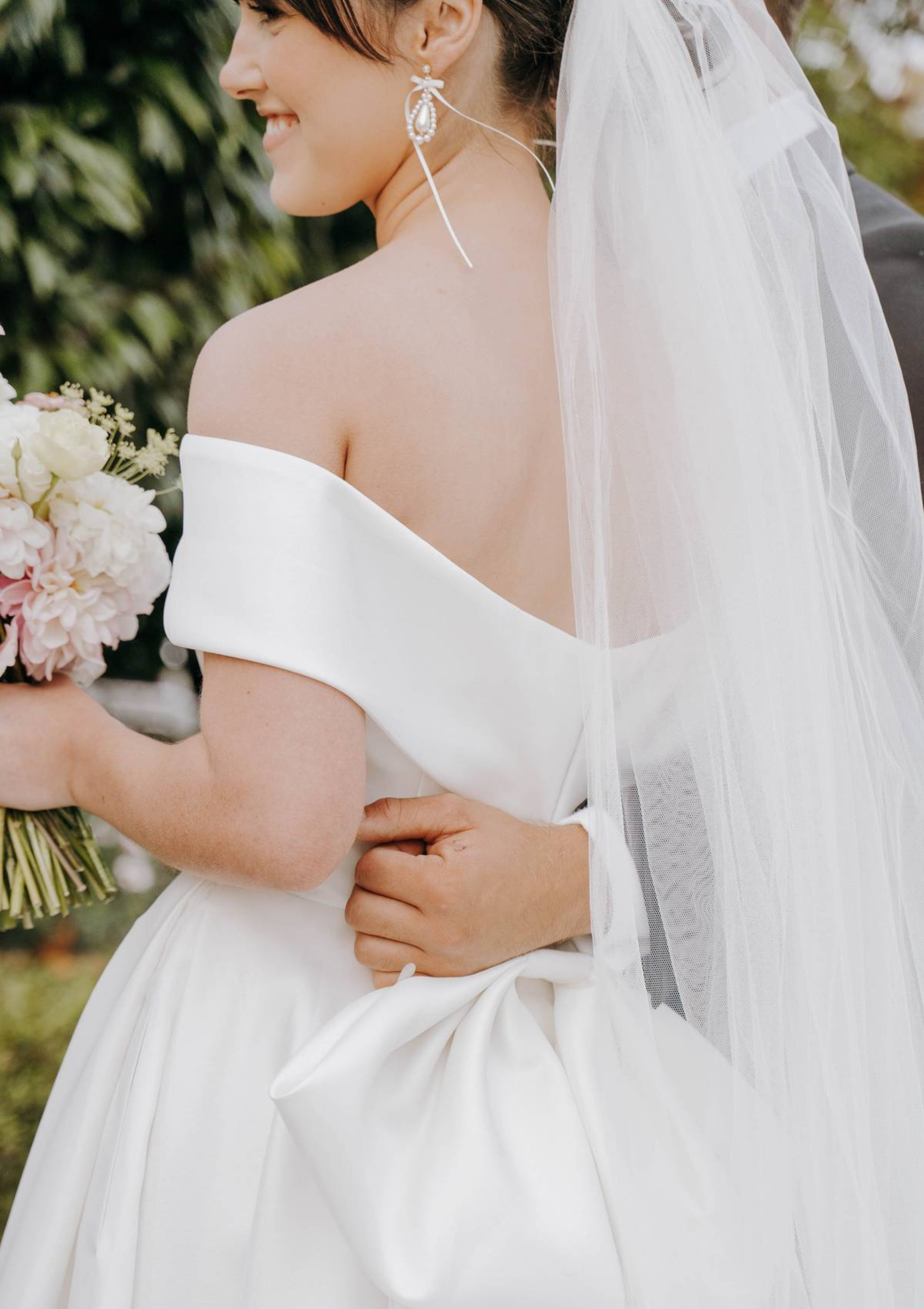 Bride in a white off-shoulder gown with a veil, holding flowers outdoors.