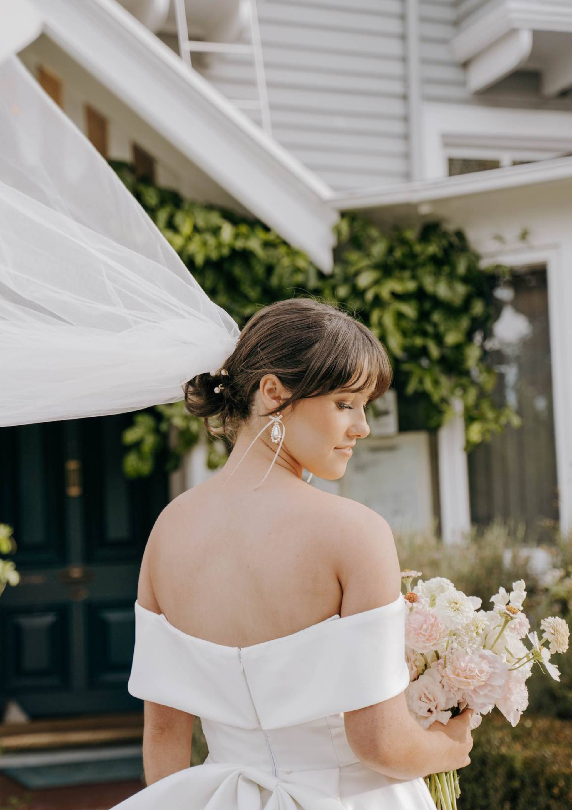 Woman in a white off-shoulder dress holding a bouquet of flowers, standing in front of a white building with greenery.