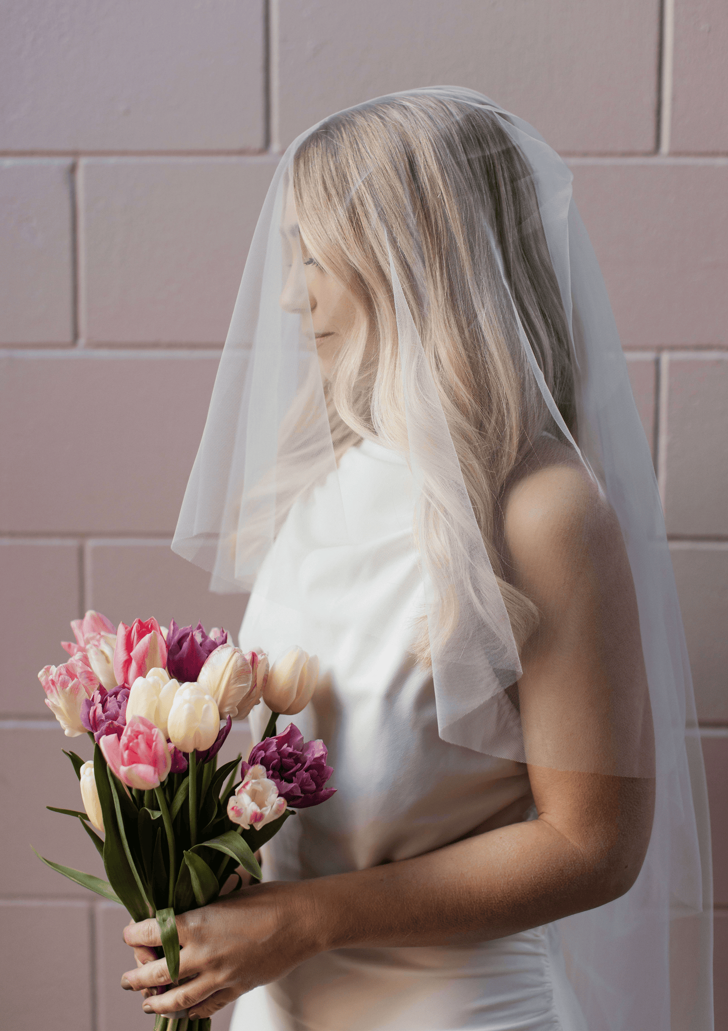 A woman wearing a classic two-tier bridal blusher veil over her face, holding a bouquet of flowers.